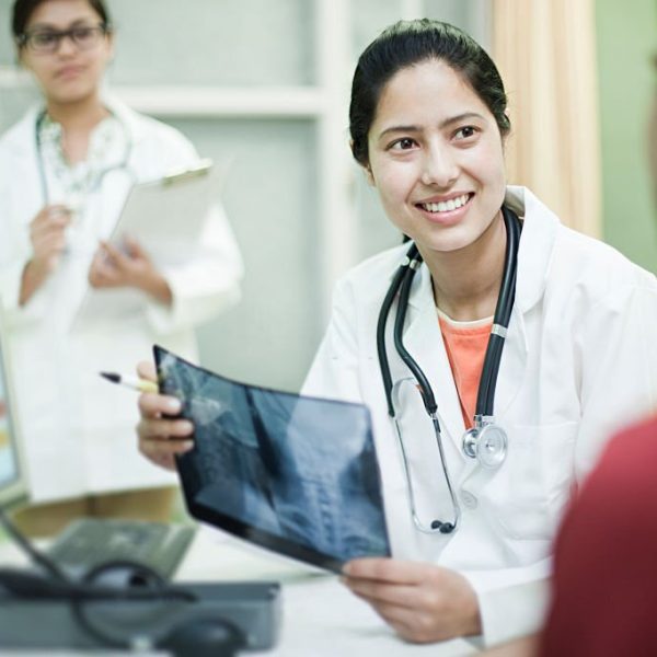 Indoor over shoulder view shot at clinic of a happy, Asian, female doctor holding an x-ray image of cervical vertebrae and talking to the male patient while junior doctor taking notes on a clipboard in the background. Three people, waist up, horizontal composition with selective focus.